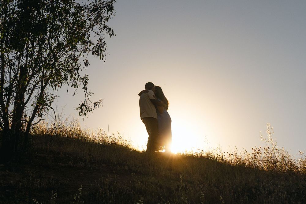 Couple embracing in silhouette against a golden sunset on a grassy hillside.