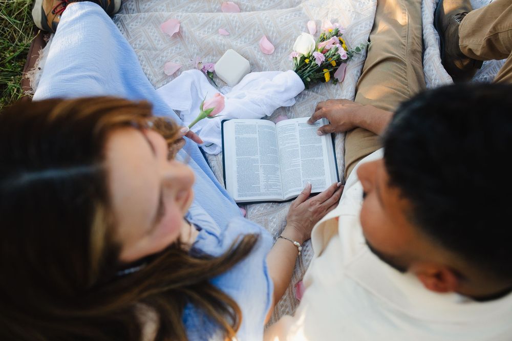 Couple reading Bible together on picnic blanket during outdoor engagement session.