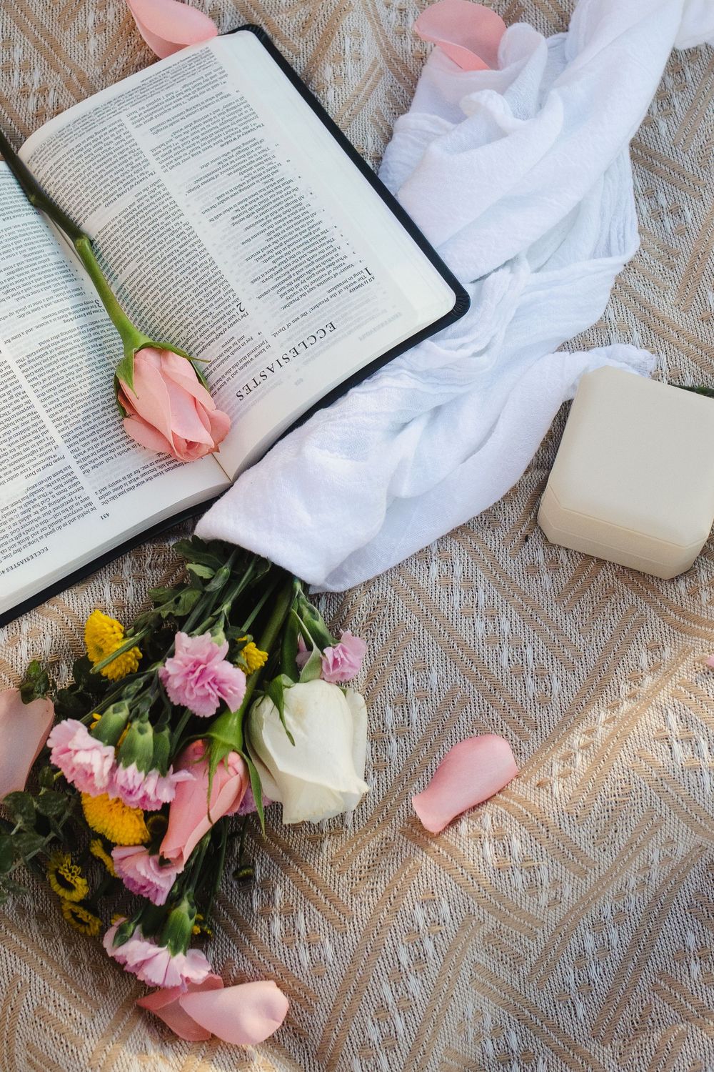 Flat lay of an open Bible, pink roses, wildflower bouquet, ring box, and white fabric on lace.