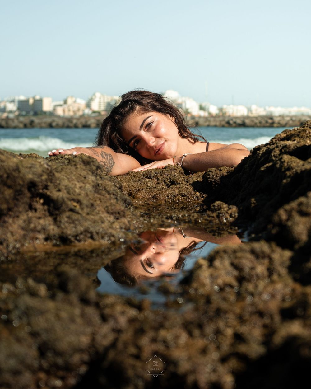 Woman with tattoo resting on rocky shore, her reflection visible in a tide pool, with a coastal city skyline in the background.