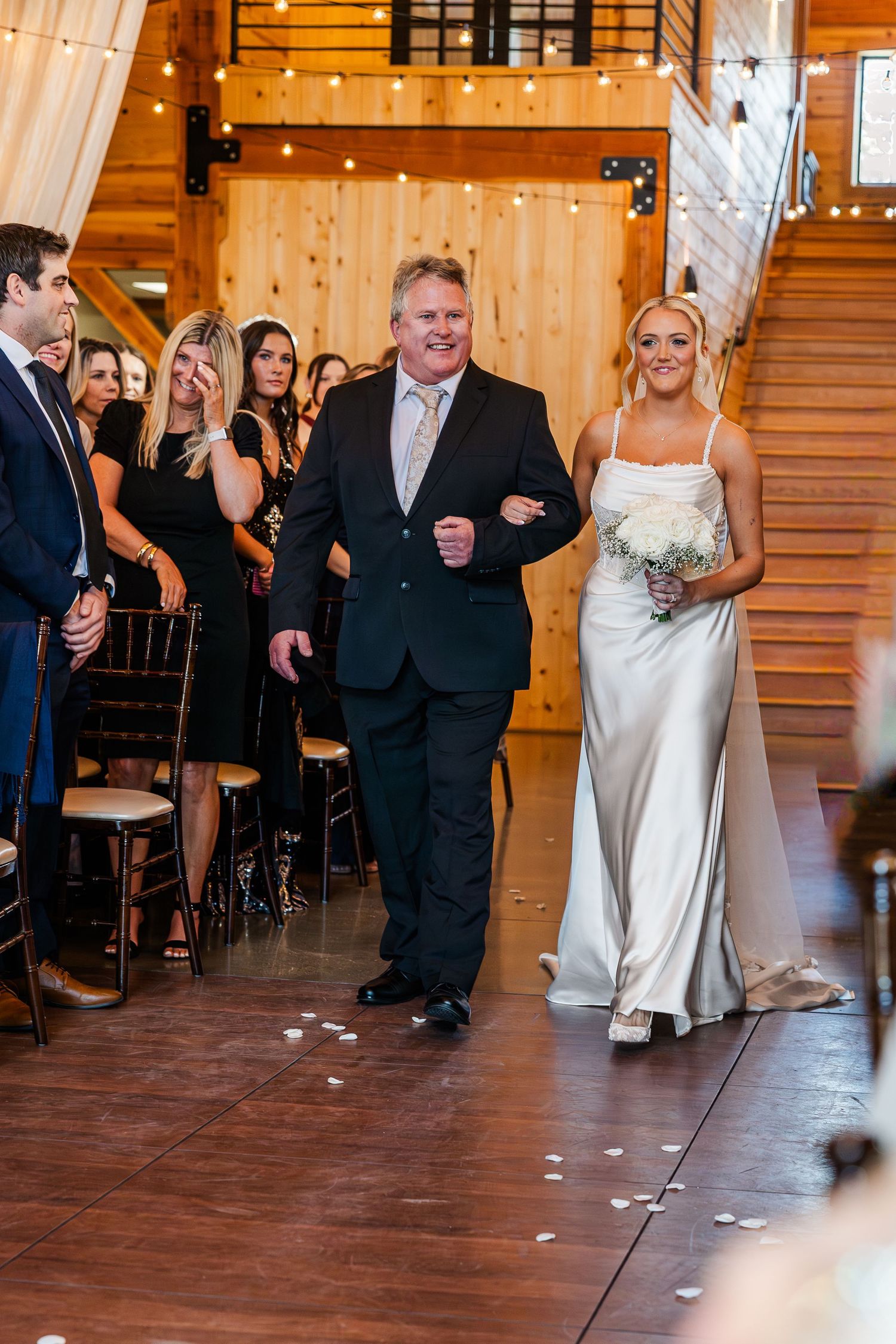 Emotional groom in beige suit wiping tears at barn wedding ceremony with groomsmen standing behind him.