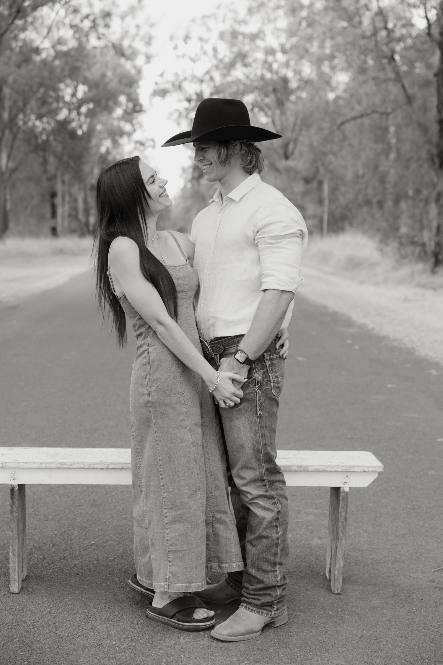 Black and white photo of a couple holding hands on a rural road, man wearing cowboy hat and boots, woman in denim dress.