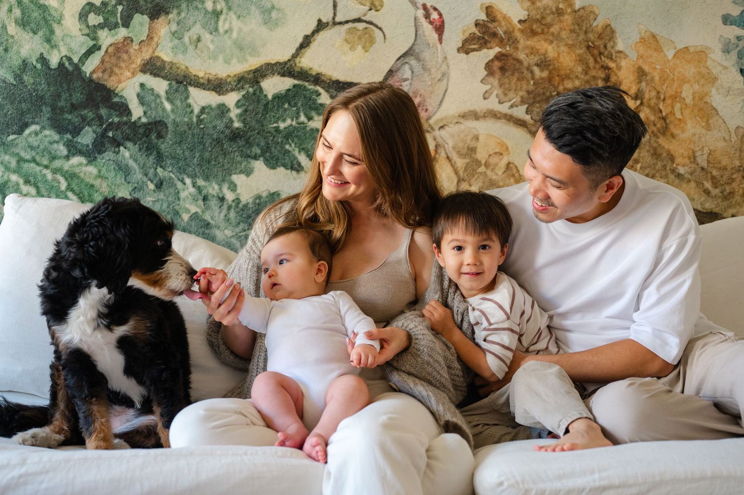Family with two young children and a dog sitting together on a white couch in front of a floral mural.