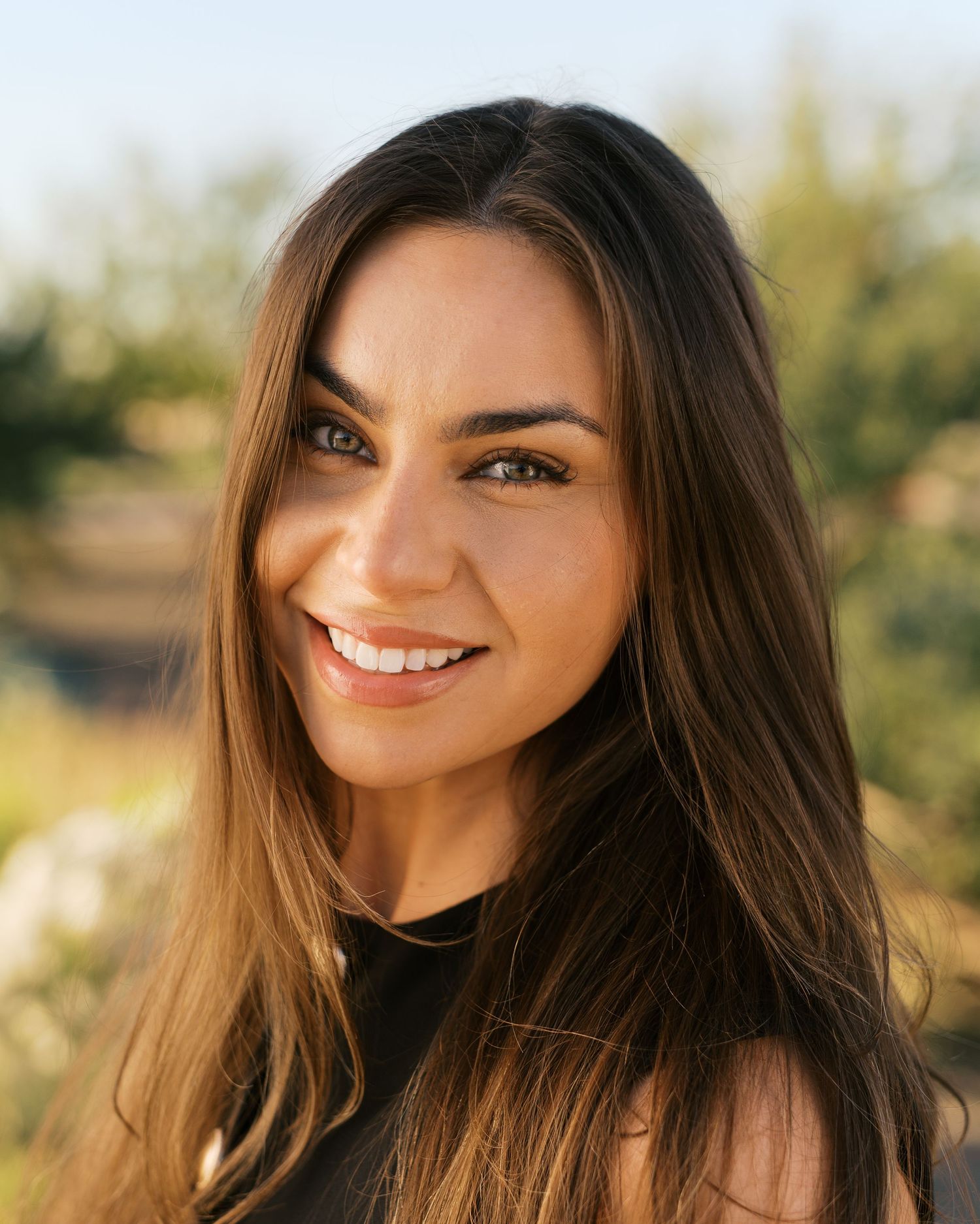 Smiling young woman with long brown hair outdoors in natural sunlight with green trees in background.