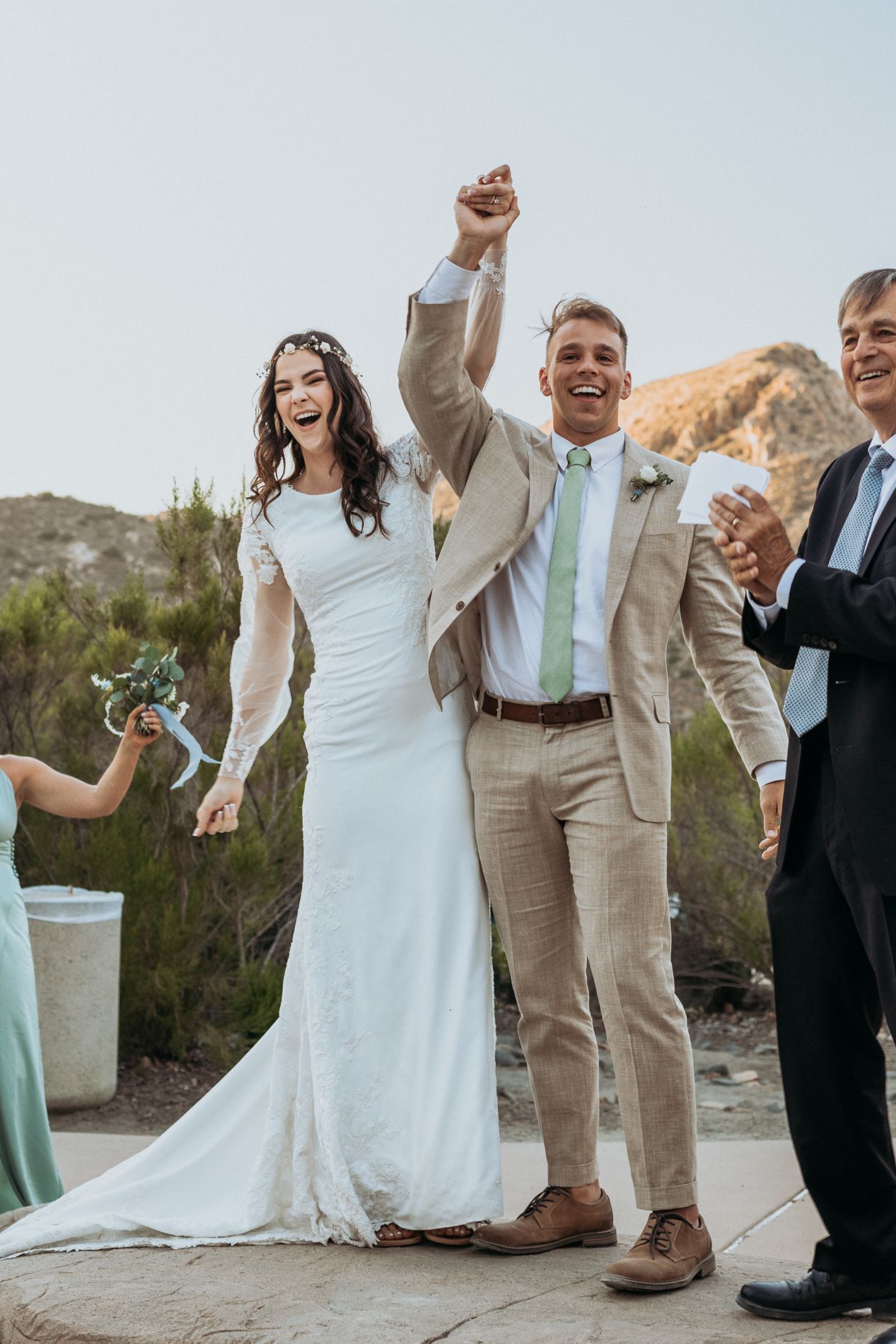 Joyful newlyweds celebrate after wedding ceremony outdoors, groom raising fist in tan suit, bride in white gown, desert backdrop.