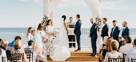 Elegant outdoor beach wedding ceremony with bride and groom at floral arch altar, guests seated, ocean backdrop.