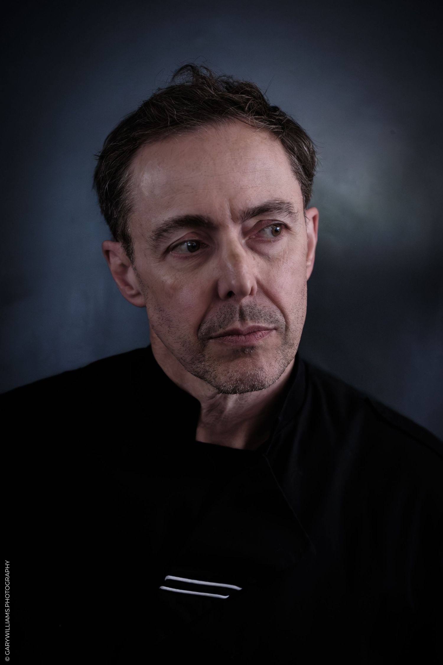 A middle-aged man with dark hair wearing a black shirt poses against a dark moody blue-gray background.