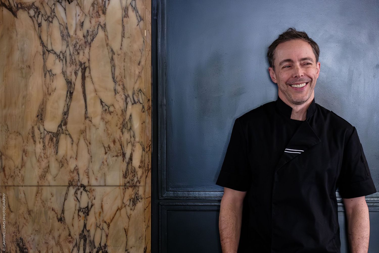 Smiling chef in black uniform standing beside marble wall in upscale restaurant setting.