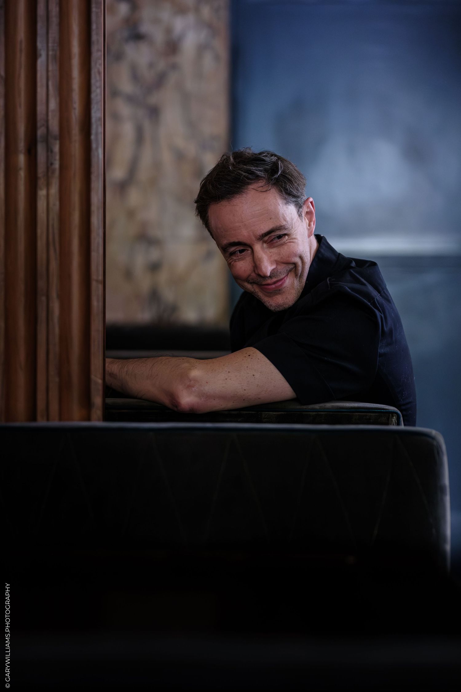 Smiling man in black shirt leaning on dark sofa in moody interior setting.