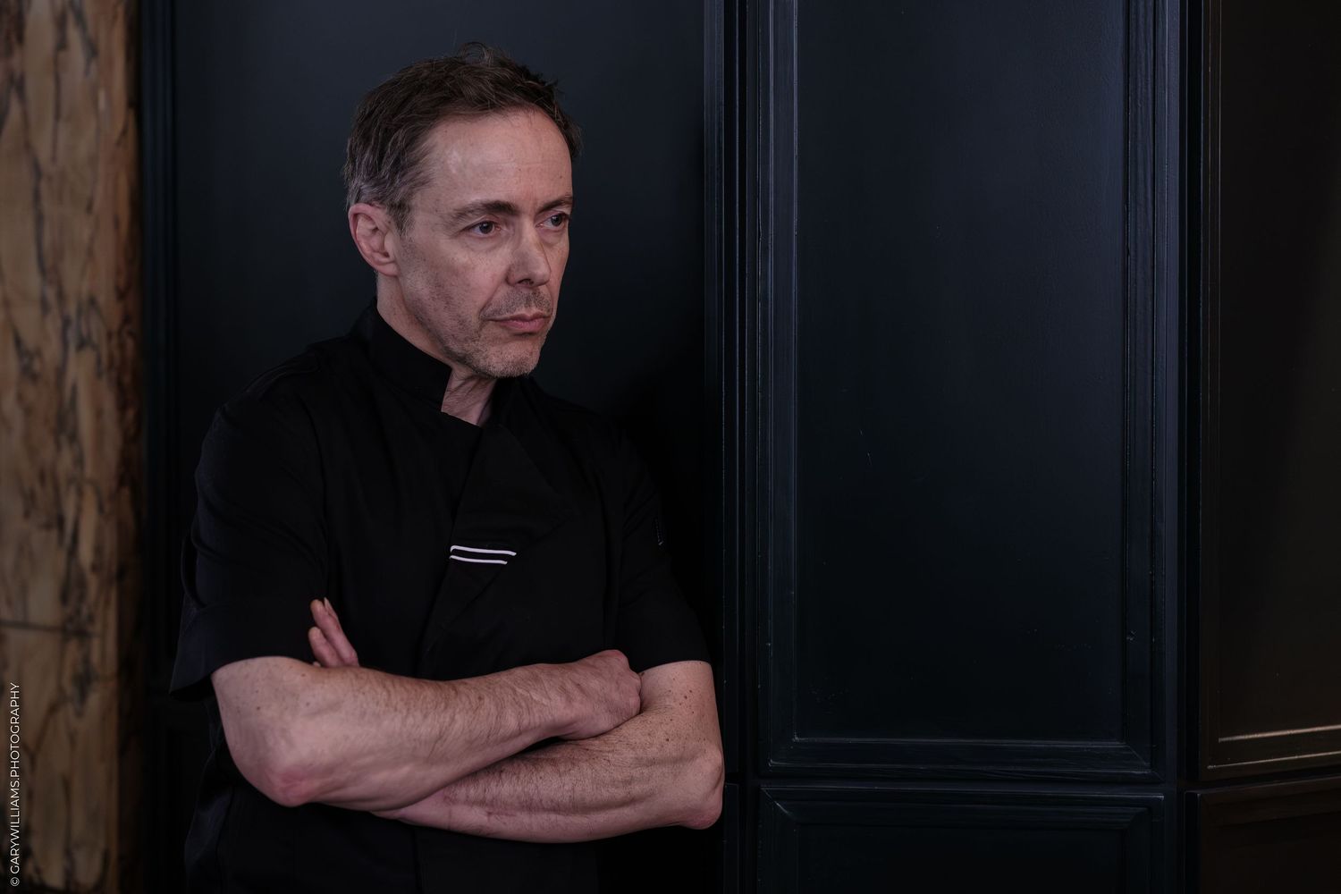 A middle-aged man in a black t-shirt stands with arms crossed against a dark paneled wall.