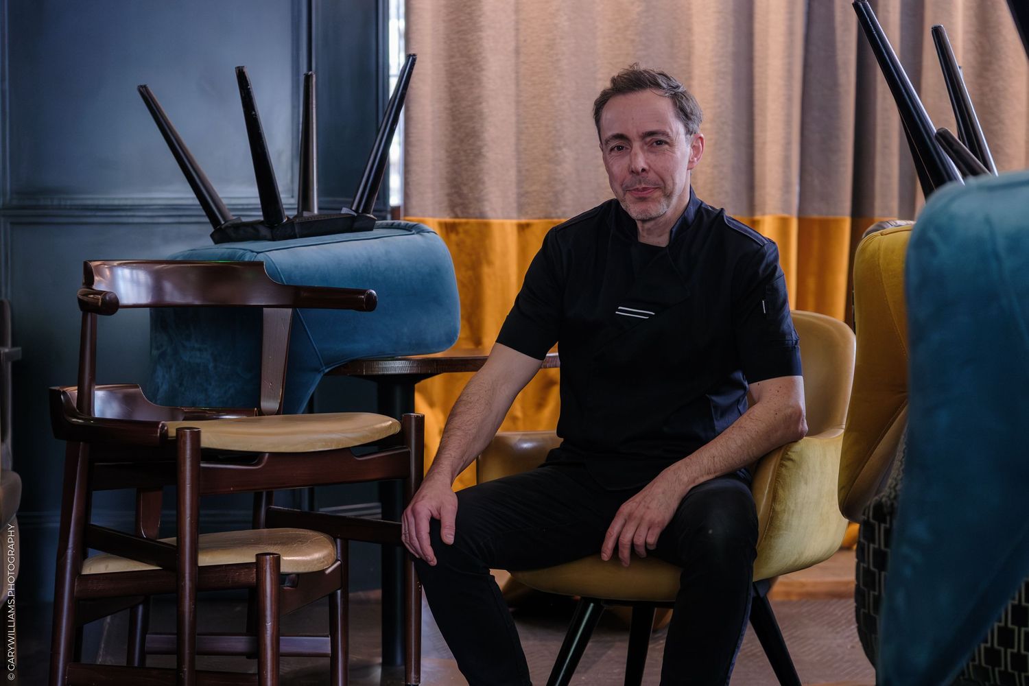 A chef in black uniform sits among vintage chairs in a stylishly decorated restaurant interior.
