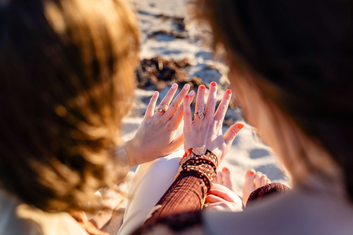 Two women comparing hands with rings and bracelets on a sunny beach, viewed from behind.