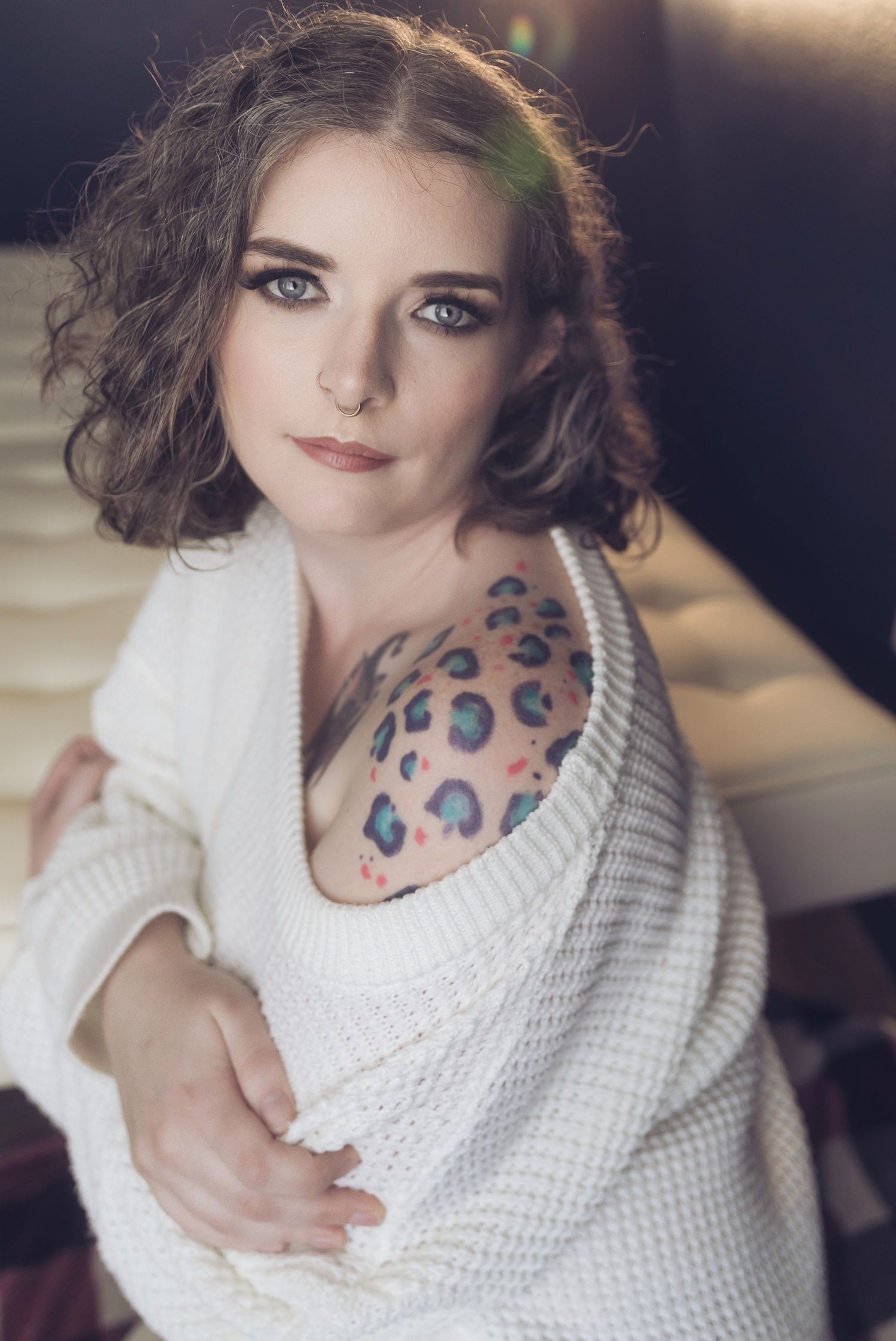 Young woman with curly hair and colorful leopard print shoulder tattoo wearing a white knit sweater.