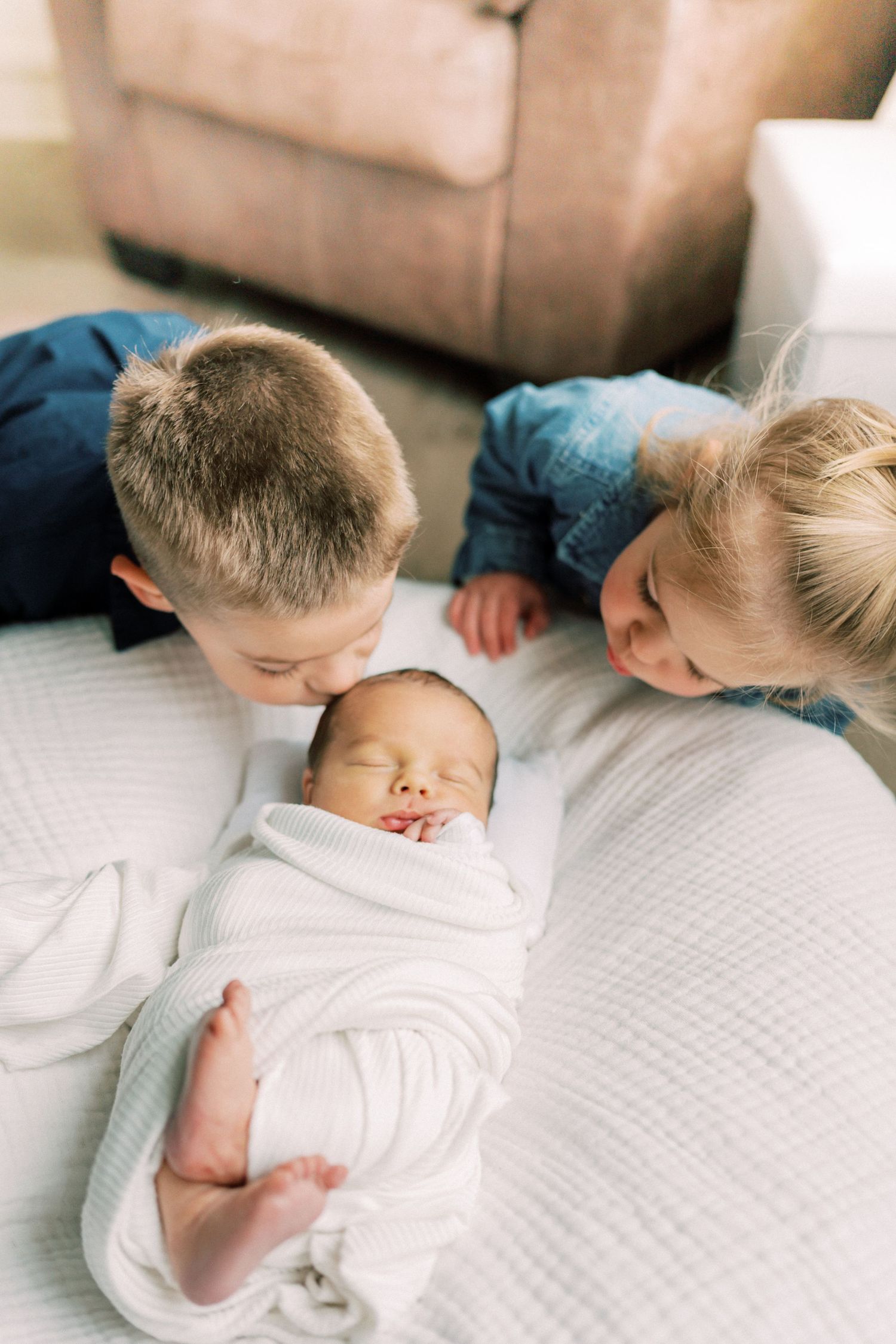 Two young siblings lovingly kiss and cuddle a swaddled newborn baby on a white bed.