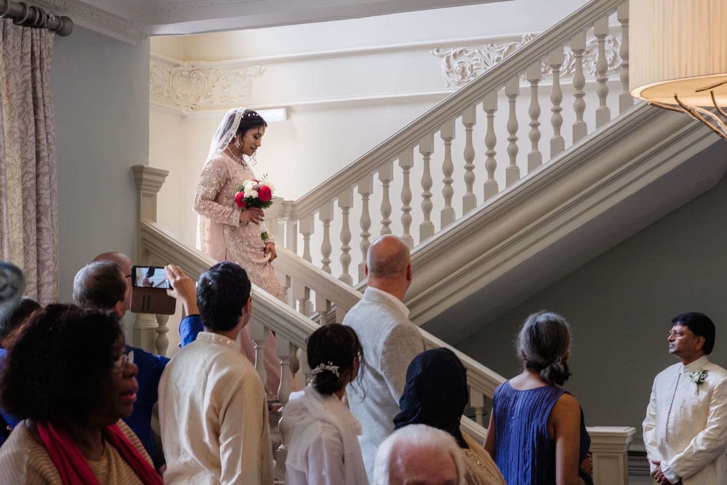 Bride descends elegant white staircase in pink gown holding red bouquet as wedding guests watch groom below.