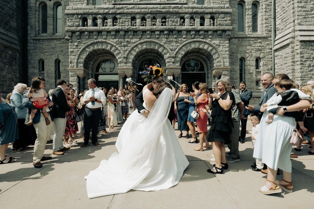Newlywed couple sharing a kiss outside a historic stone church surrounded by wedding guests on a sunny day.