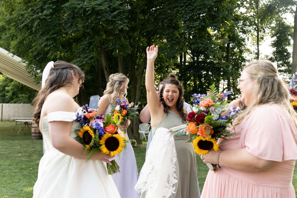 Joyful bride celebrates with bridesmaids holding colorful sunflower bouquets outdoors at a wedding reception.