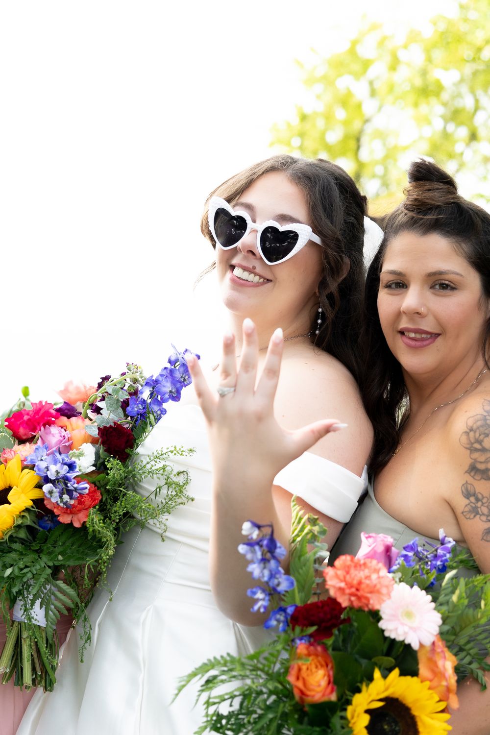 Two brides in white dresses holding colorful bouquets, one wearing heart sunglasses showing wedding ring.