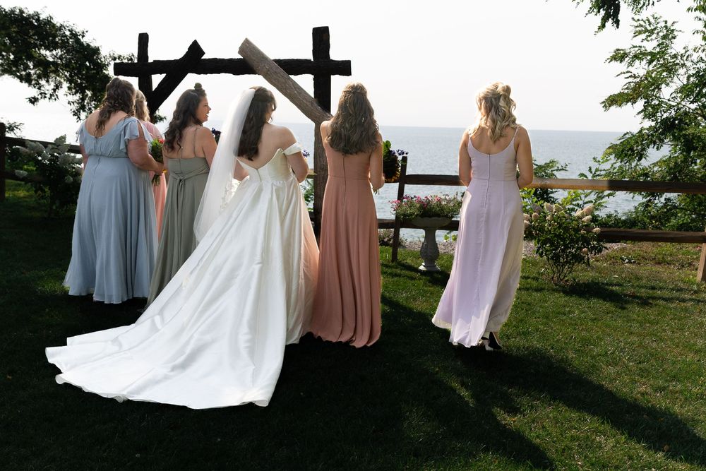 Bride and bridesmaids in mismatched gowns stand before rustic arch overlooking scenic waterfront.