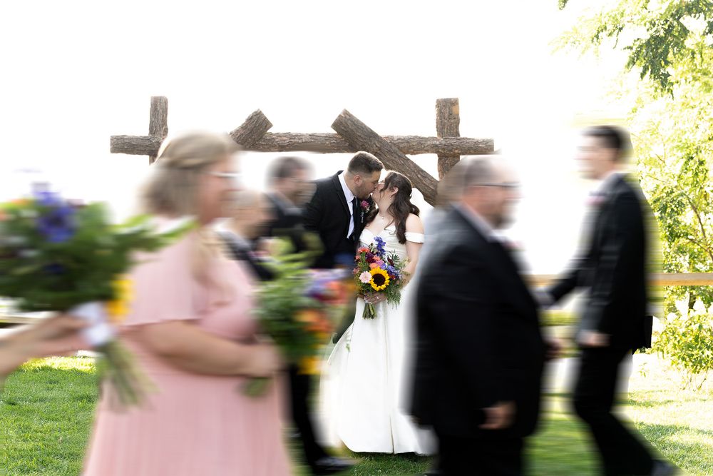 Bride and groom kissing under rustic wooden arch at outdoor wedding ceremony with bridal party in motion.