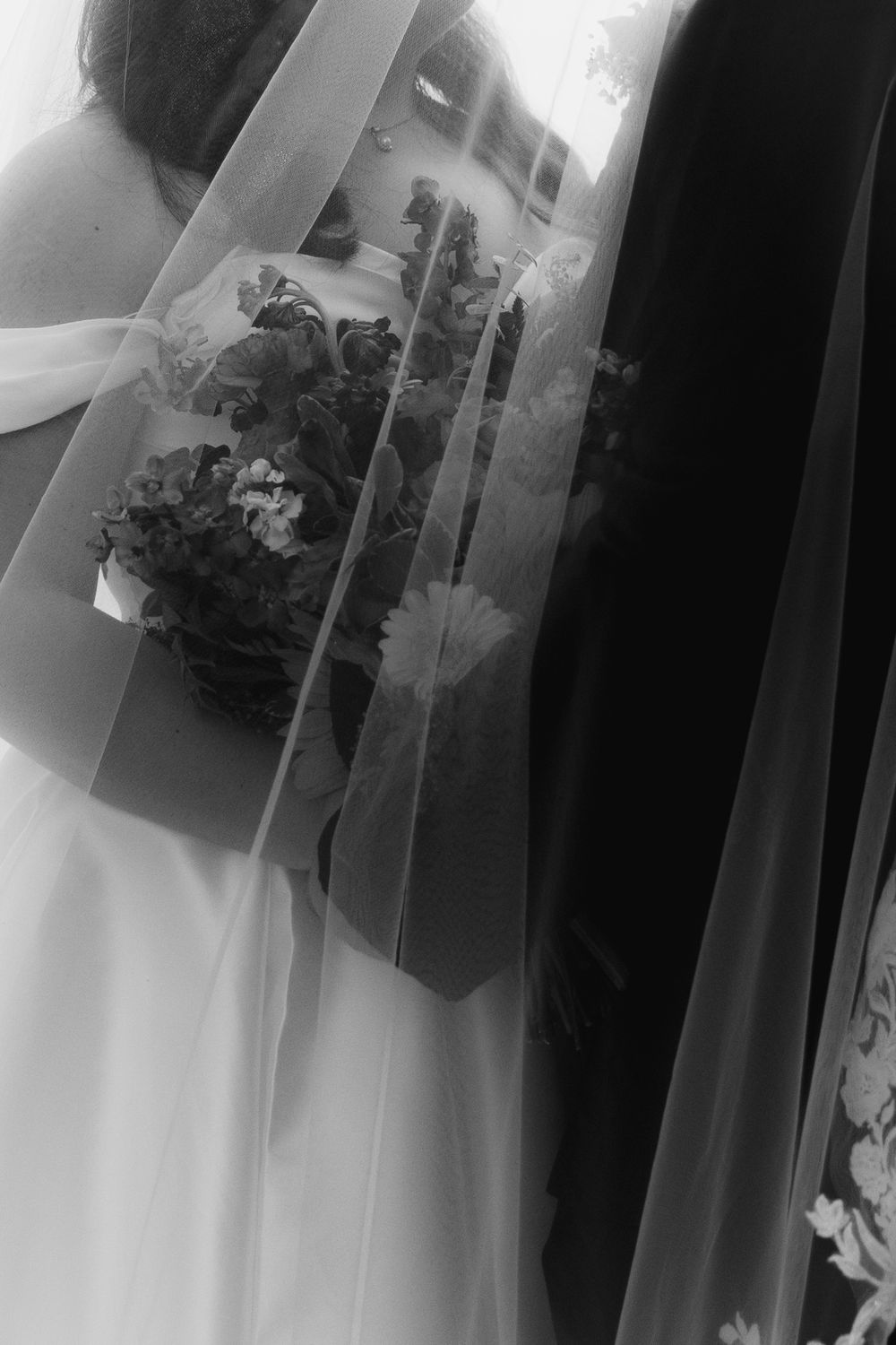 Black and white artistic photo of bride holding floral bouquet through flowing wedding veil.