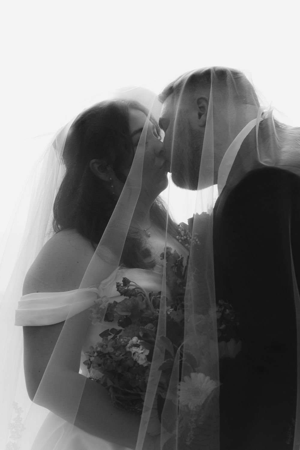 Romantic black and white photo of bride and groom kissing under flowing wedding veil.
