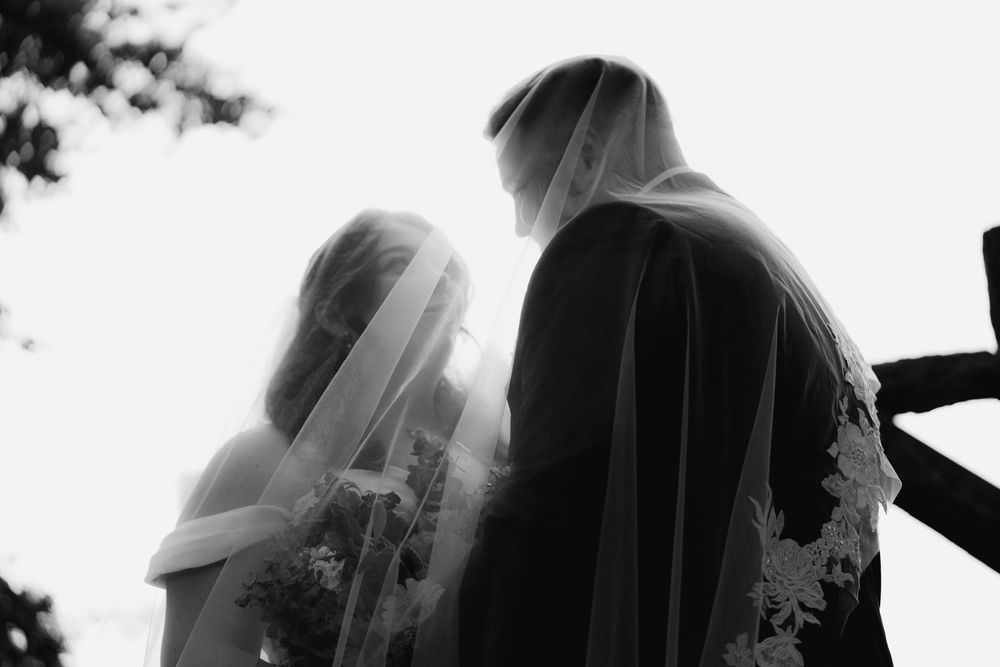 Black and white silhouette of bride and groom under wedding veil during outdoor ceremony.