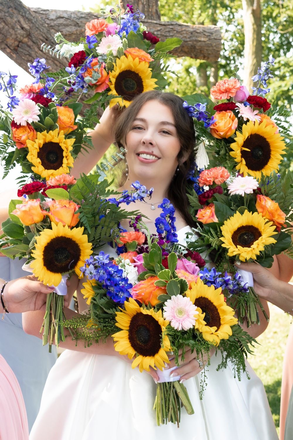 Smiling bride surrounded by colorful bouquets featuring sunflowers, roses, and blue flowers at outdoor wedding.