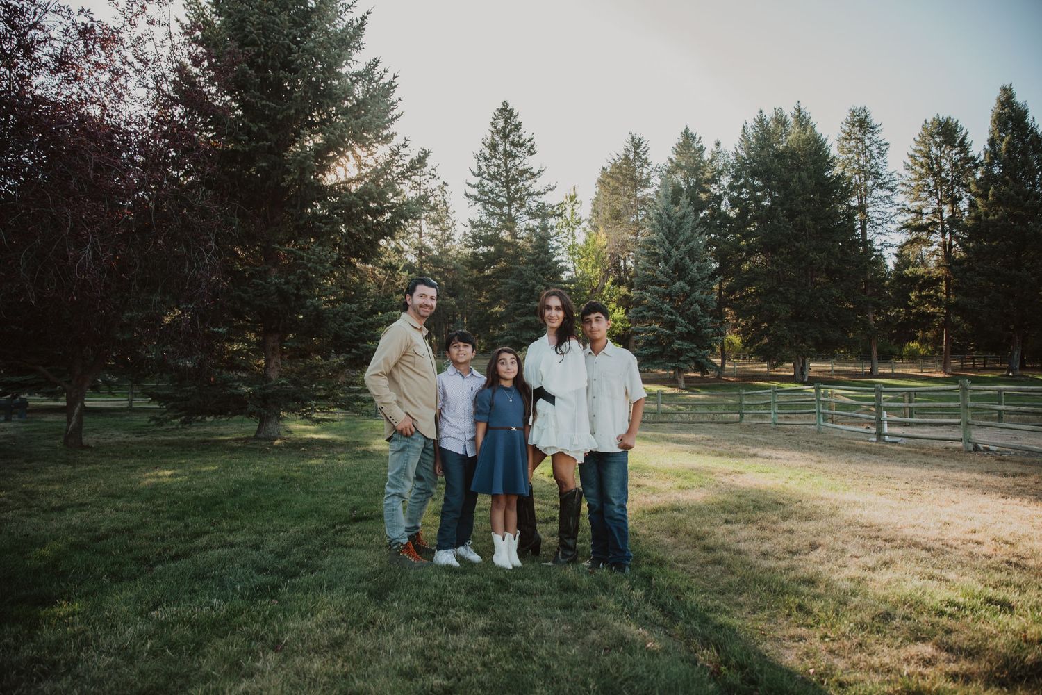 Family of five posing together on green grass surrounded by tall trees during a sunny outdoor photo session.