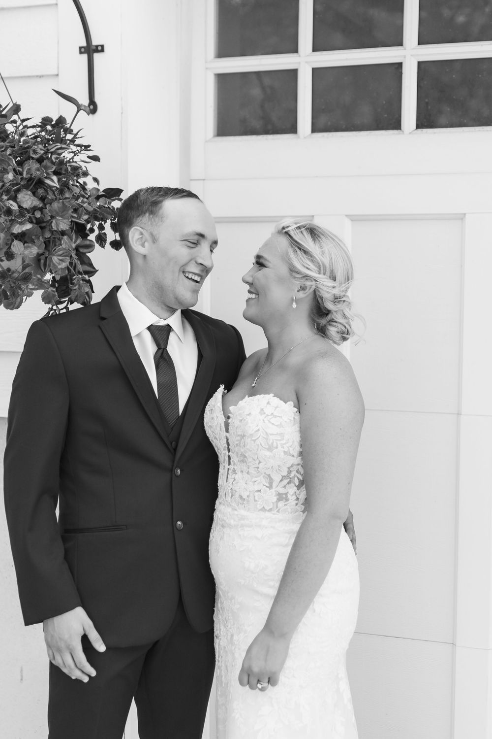 Black and white wedding photo of smiling bride in lace gown and groom in dark suit standing together outdoors.