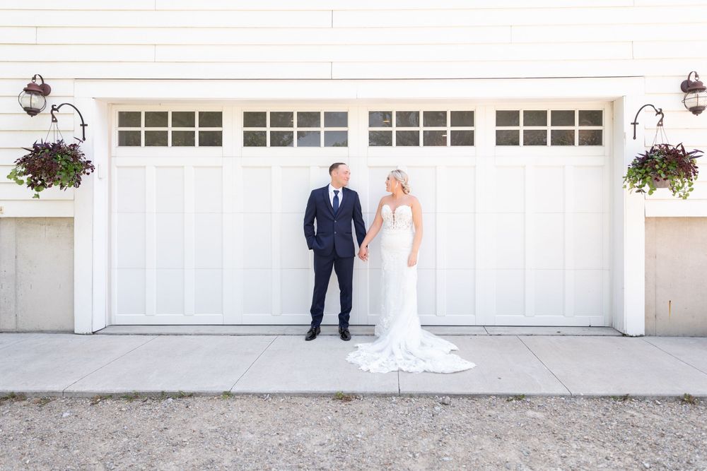 Bride and groom standing apart in front of white barn doors with hanging flower baskets on their wedding day.