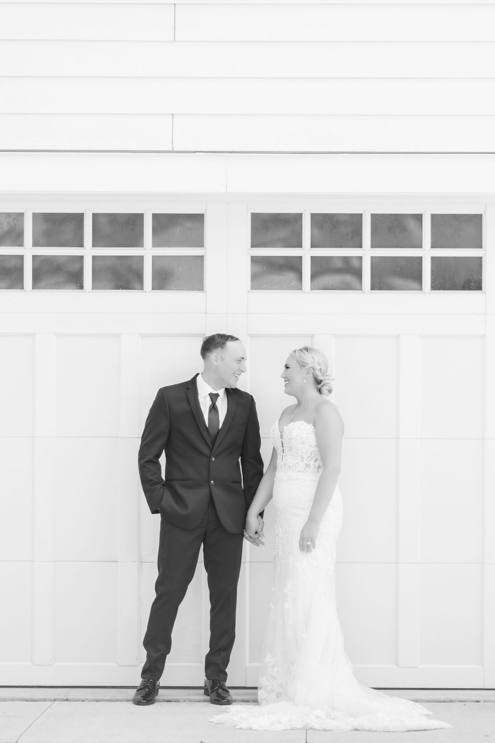 Black and white photo of a bride and groom holding hands, smiling at each other in front of a white garage door.