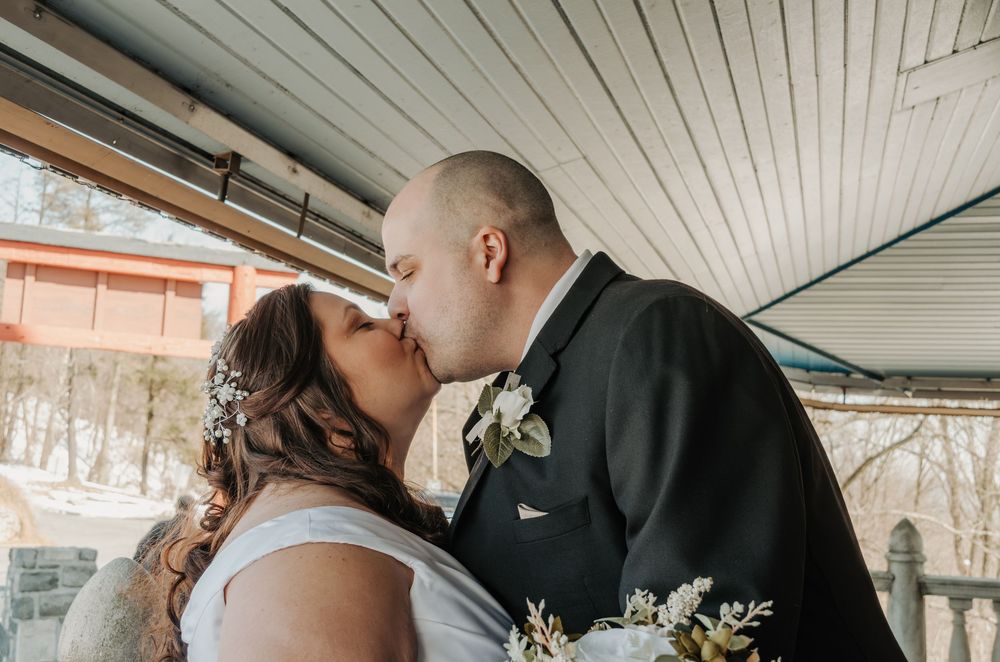 Bride and groom share a first kiss on a porch, bride holding white floral bouquet with greenery on their wedding day.