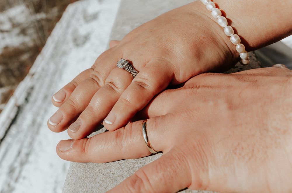 Two hands displaying wedding rings, one with a pearl bracelet, resting together on a stone surface.