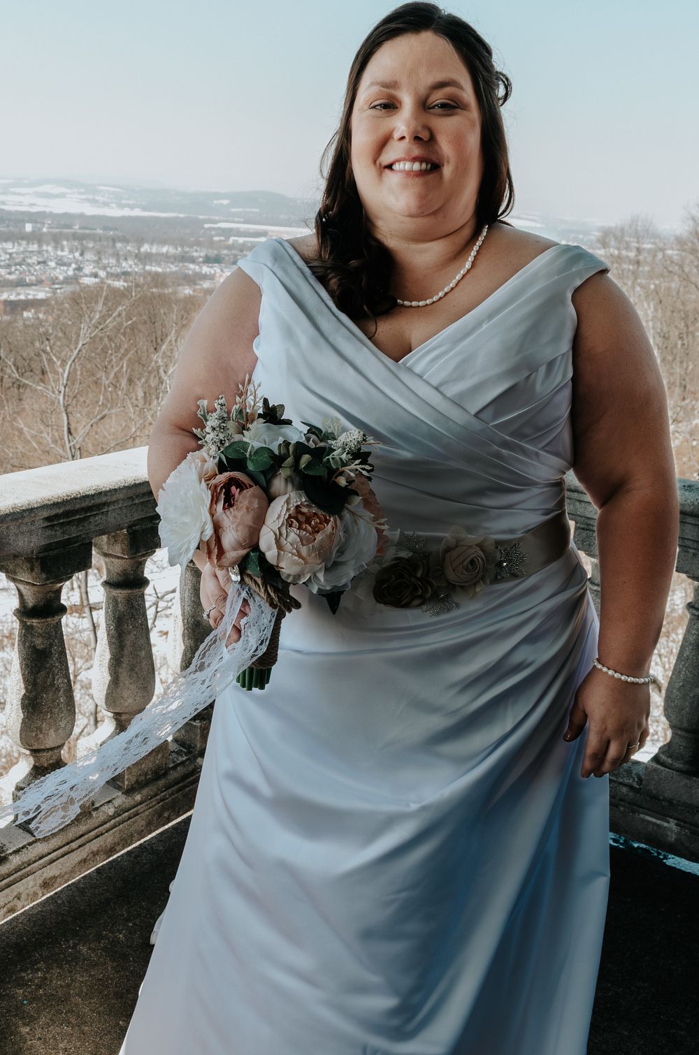 Smiling bride in light blue gown holding floral bouquet on stone balcony with winter landscape backdrop.