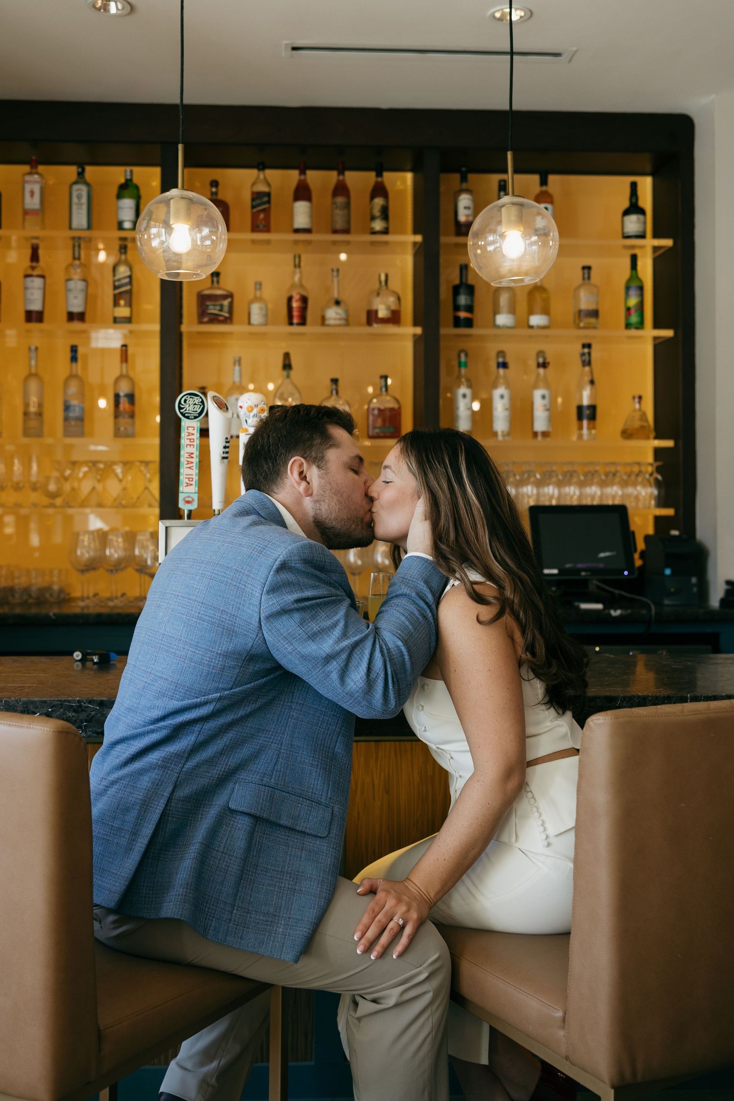 Couple sharing a romantic kiss at a stylish bar with backlit liquor shelves and pendant lights.