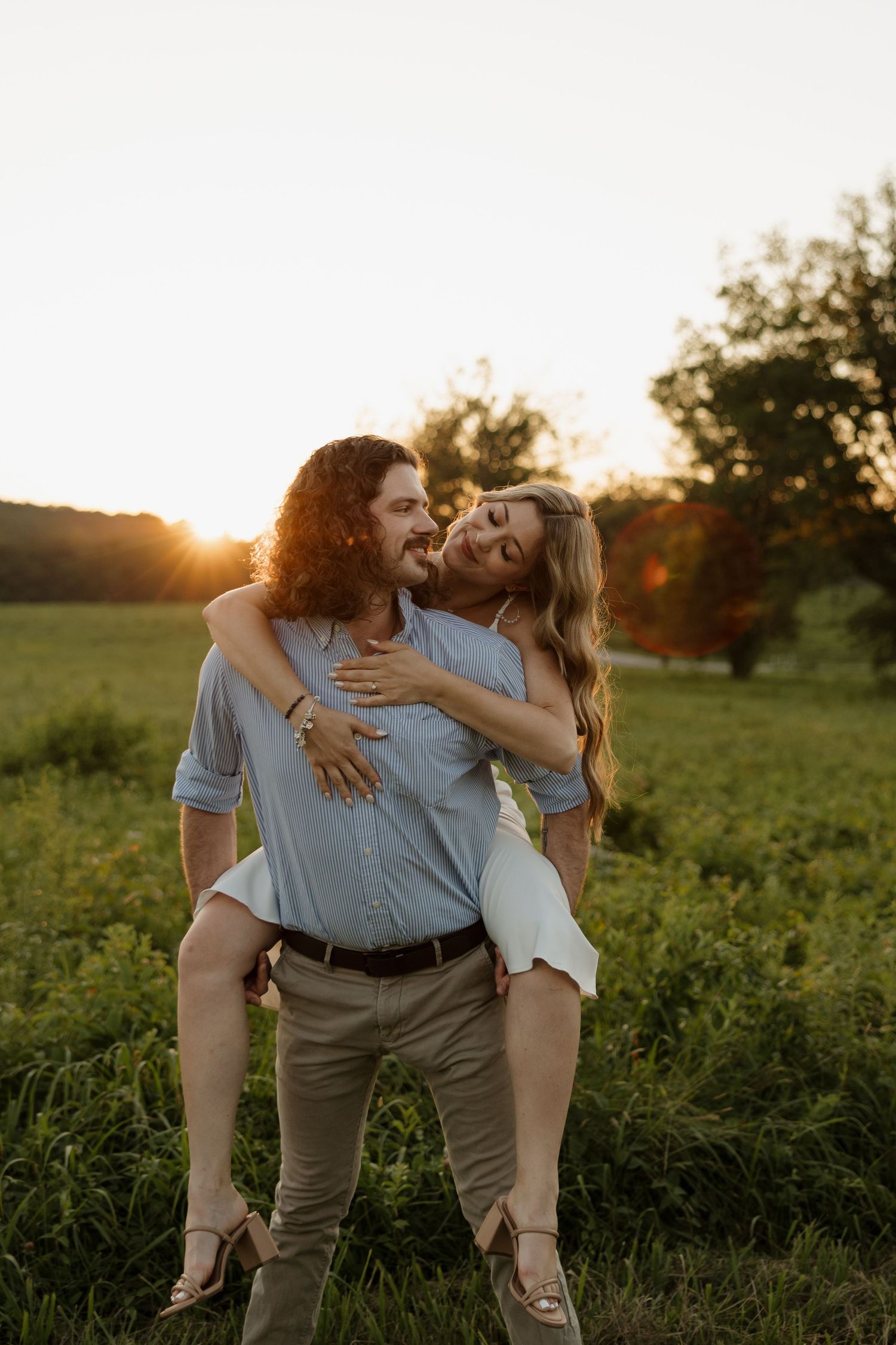 Couple sharing a romantic piggyback ride in a sunlit green field at golden hour, woman laughing joyfully.