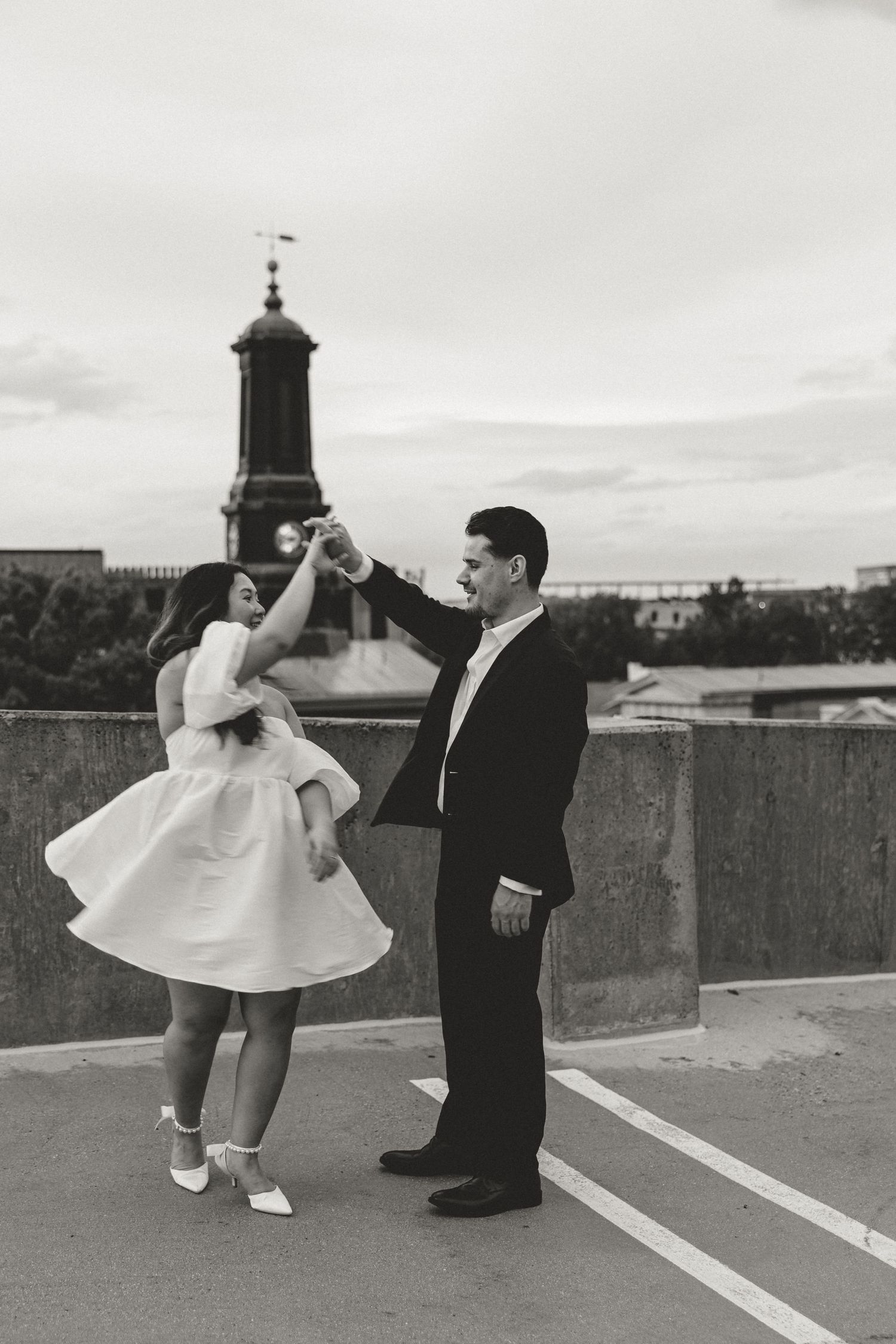 Black and white photo of a couple dancing on a rooftop, man in suit spinning woman in white dress, church tower background.