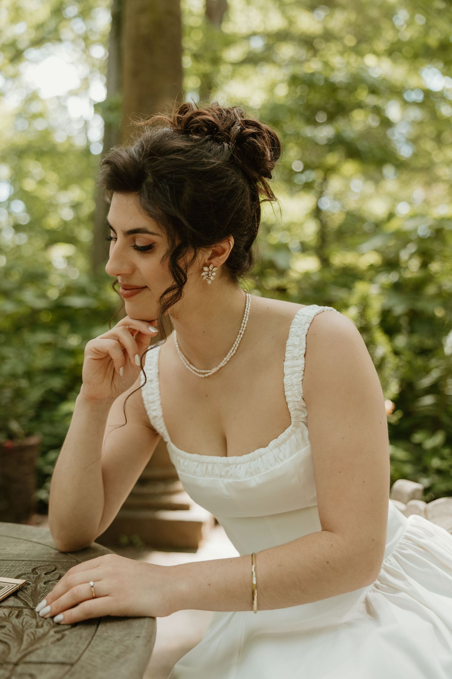 Elegant bride in white dress with pearl necklace and earrings, seated outdoors in a lush green garden setting.
