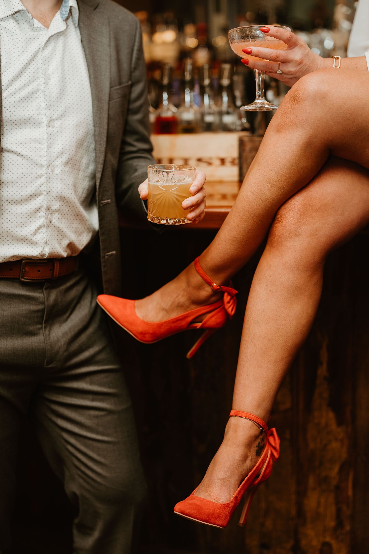 Couple at a bar holding cocktails, woman sitting with red suede heels, man in gray suit with whiskey glass.