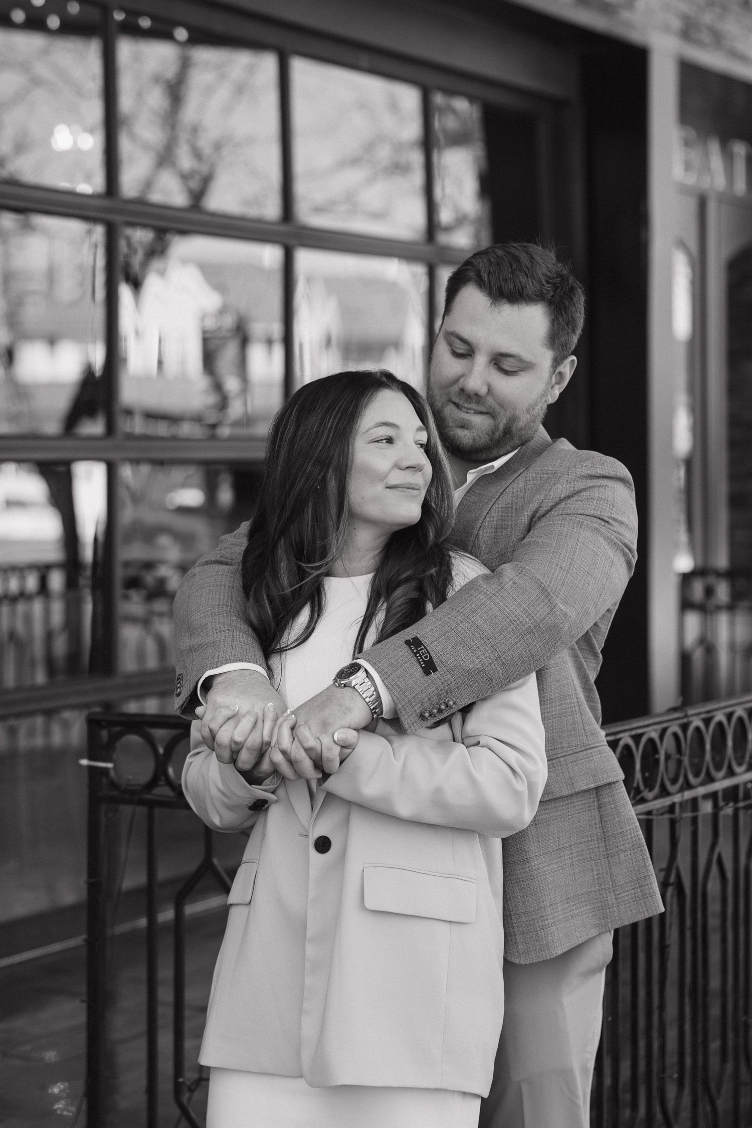 Black and white photo of a couple embracing outdoors, man in suit jacket holding woman from behind, both smiling warmly.