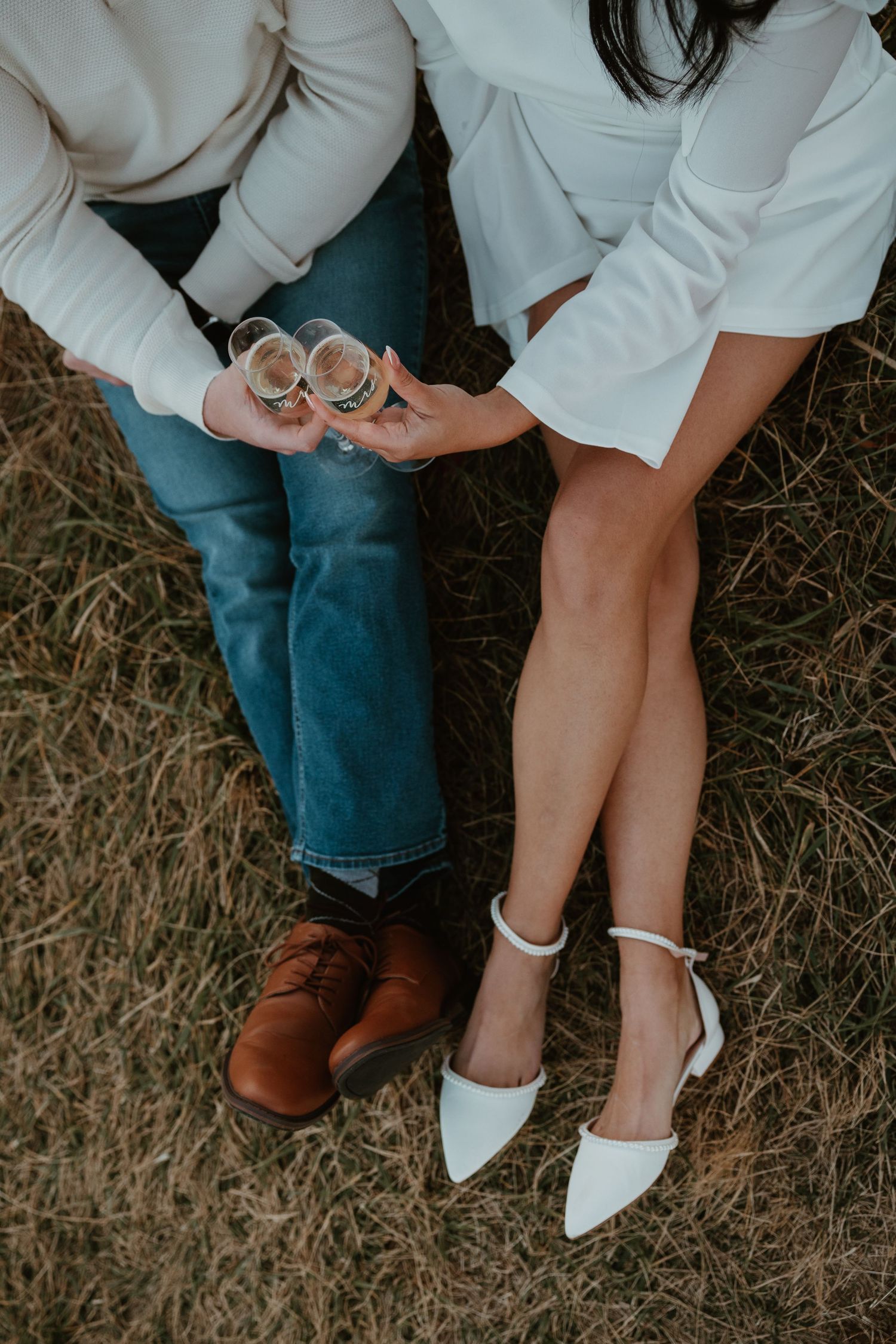 Couple sitting on grass clinking champagne glasses, wearing white outfits, blue jeans, brown leather shoes and white heels.