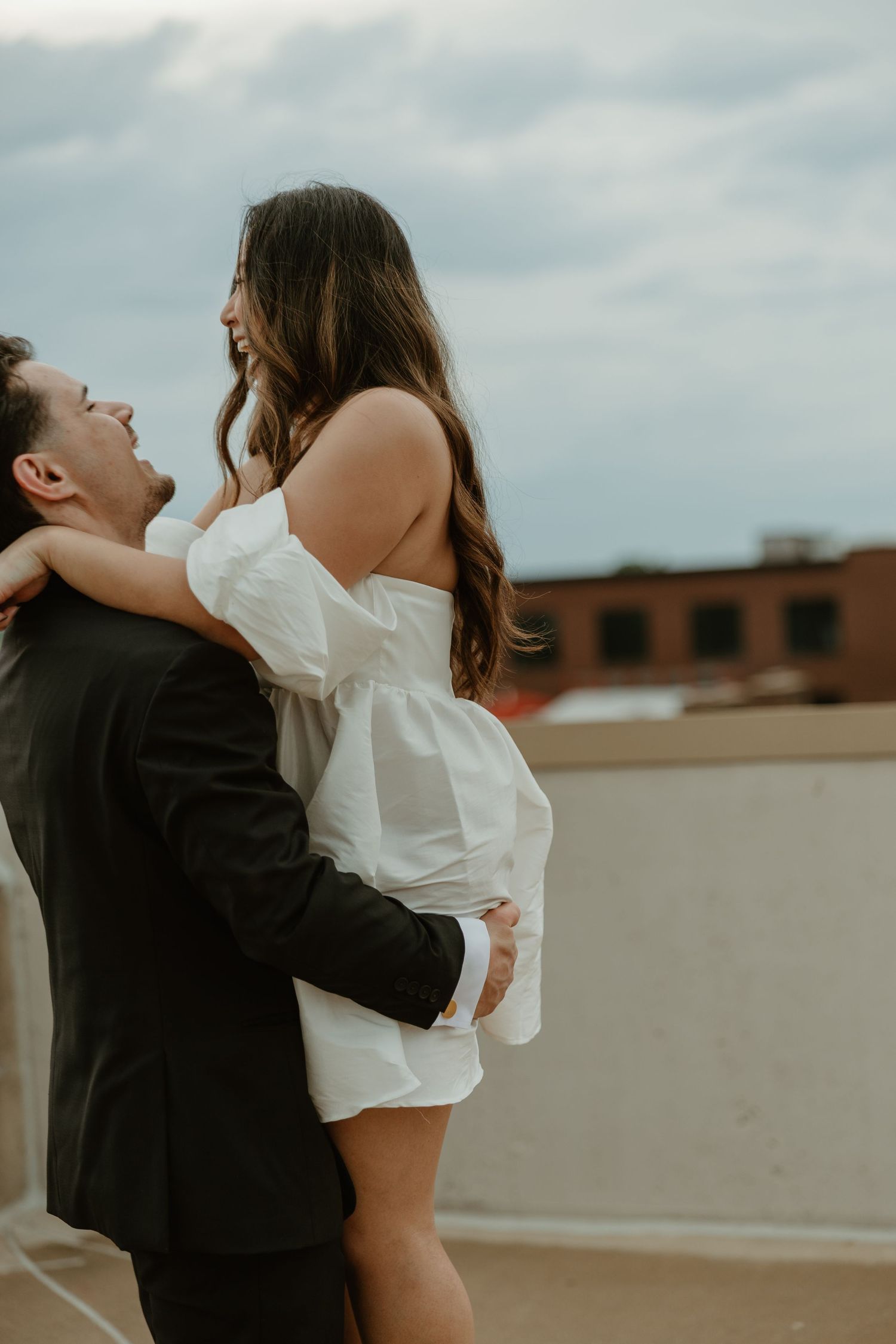 A couple embraces on a rooftop, the woman in a white off-shoulder mini dress laughing joyfully as the man holds her close.