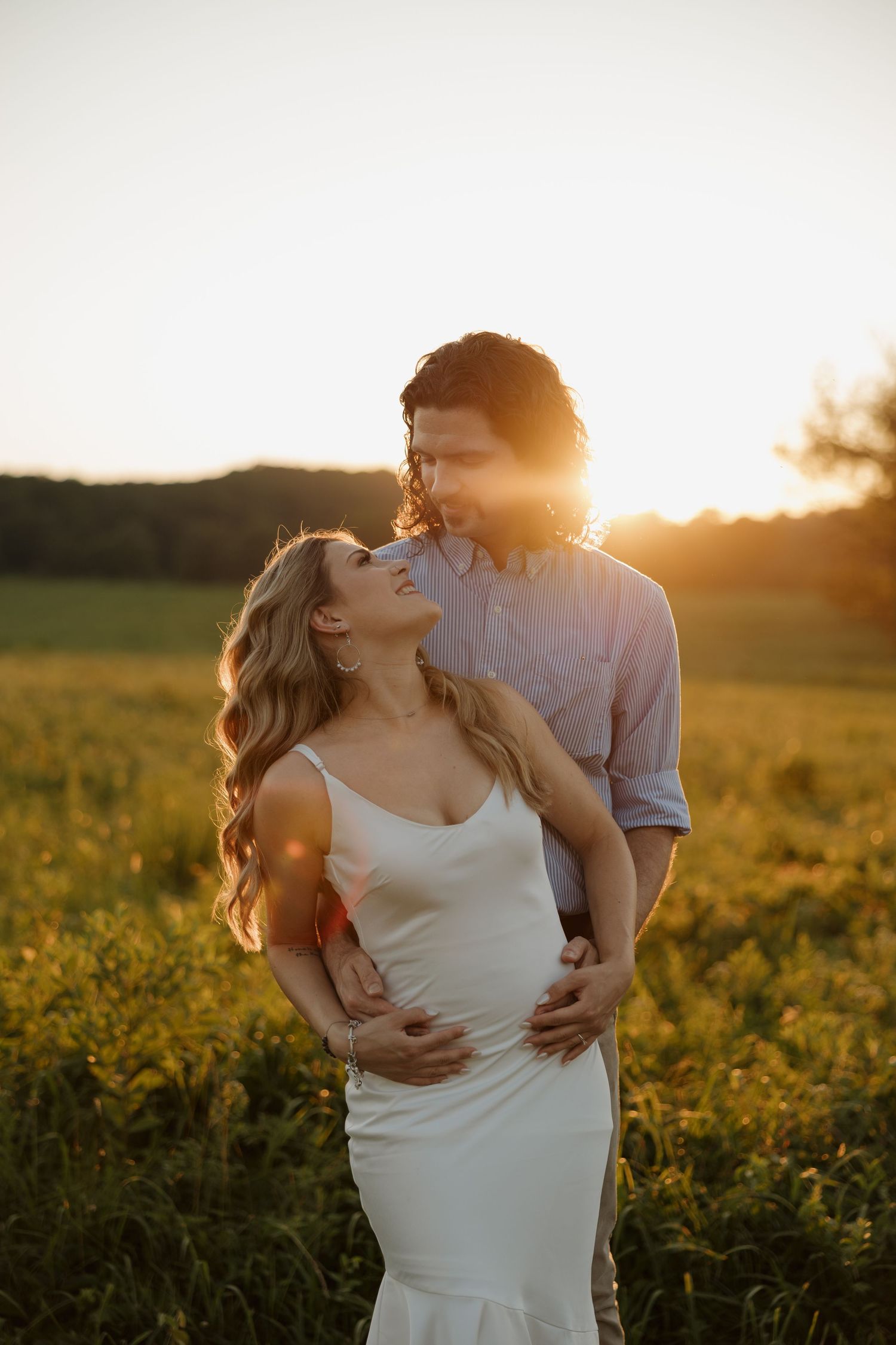 Couple embracing in golden sunset field, pregnant woman in white dress smiling up at partner during maternity photo session.