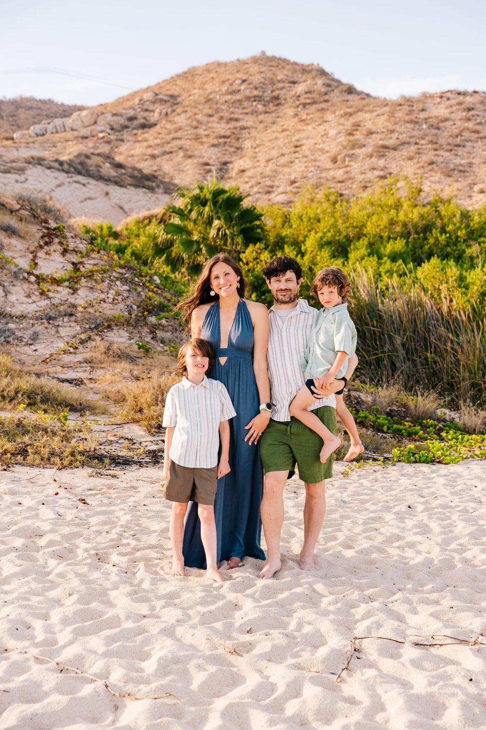 Family of four smiling on sandy beach with green hills backdrop, parents and two young boys in coordinated outfits.