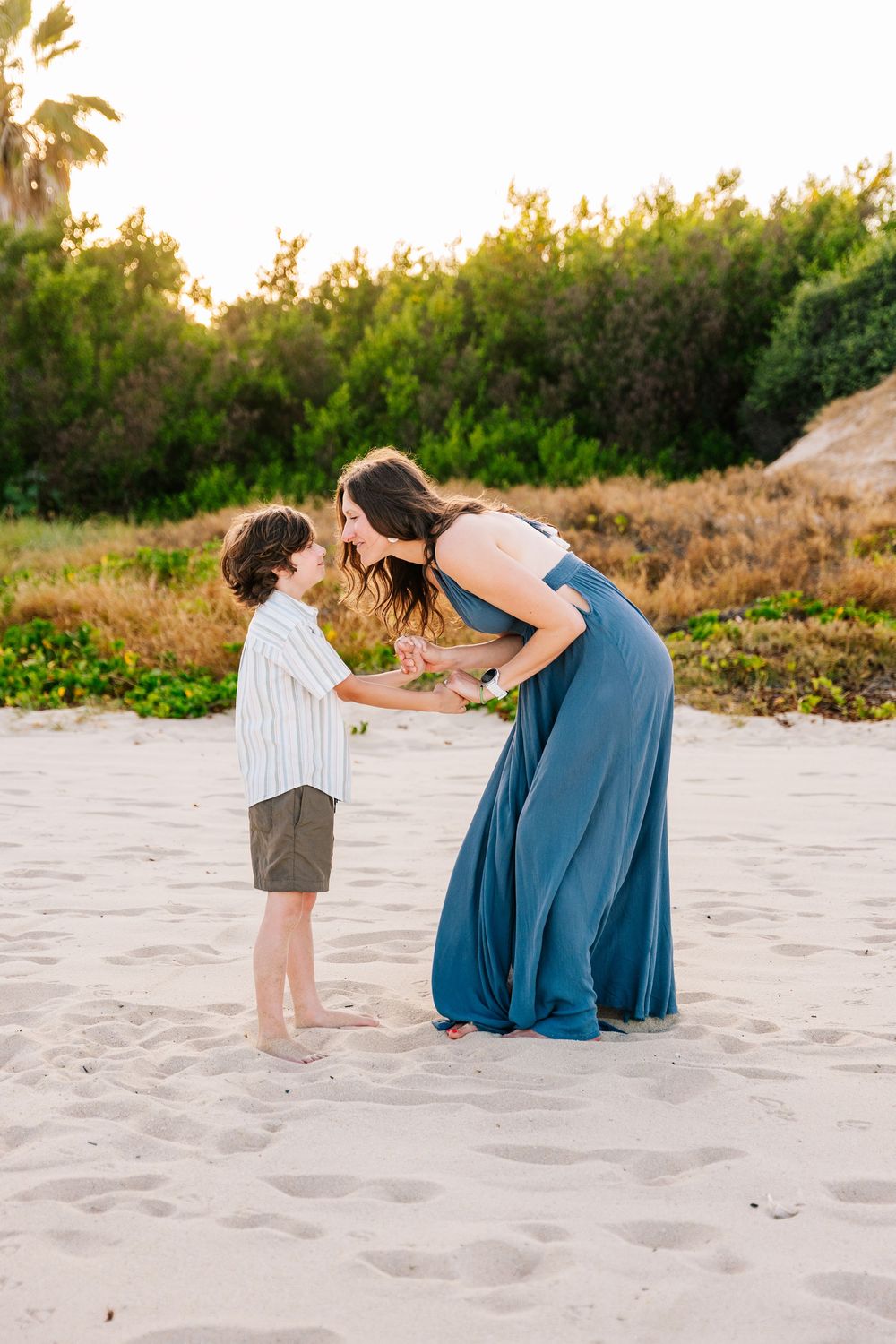 A mother in a blue dress bends down to interact with her young son on a sandy beach at golden hour.