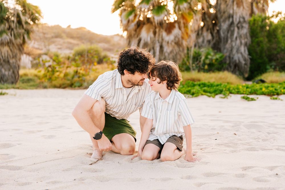 A father and young son sit together on a sandy beach, sharing a tender moment surrounded by palm trees.