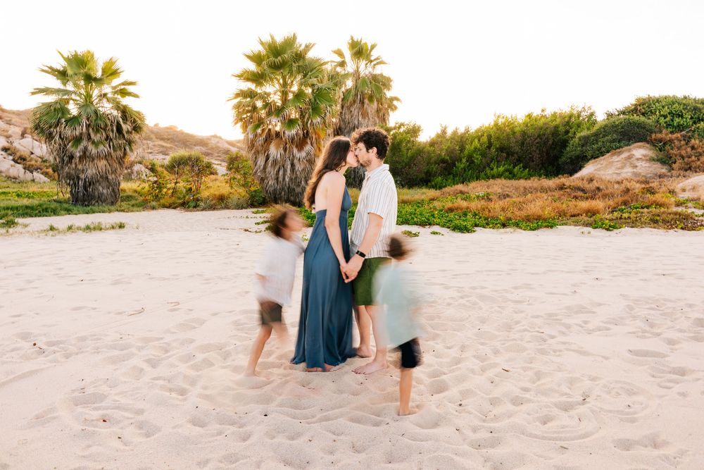 Couple kissing on sandy beach with palm trees, children playing around them in motion blur at golden hour.