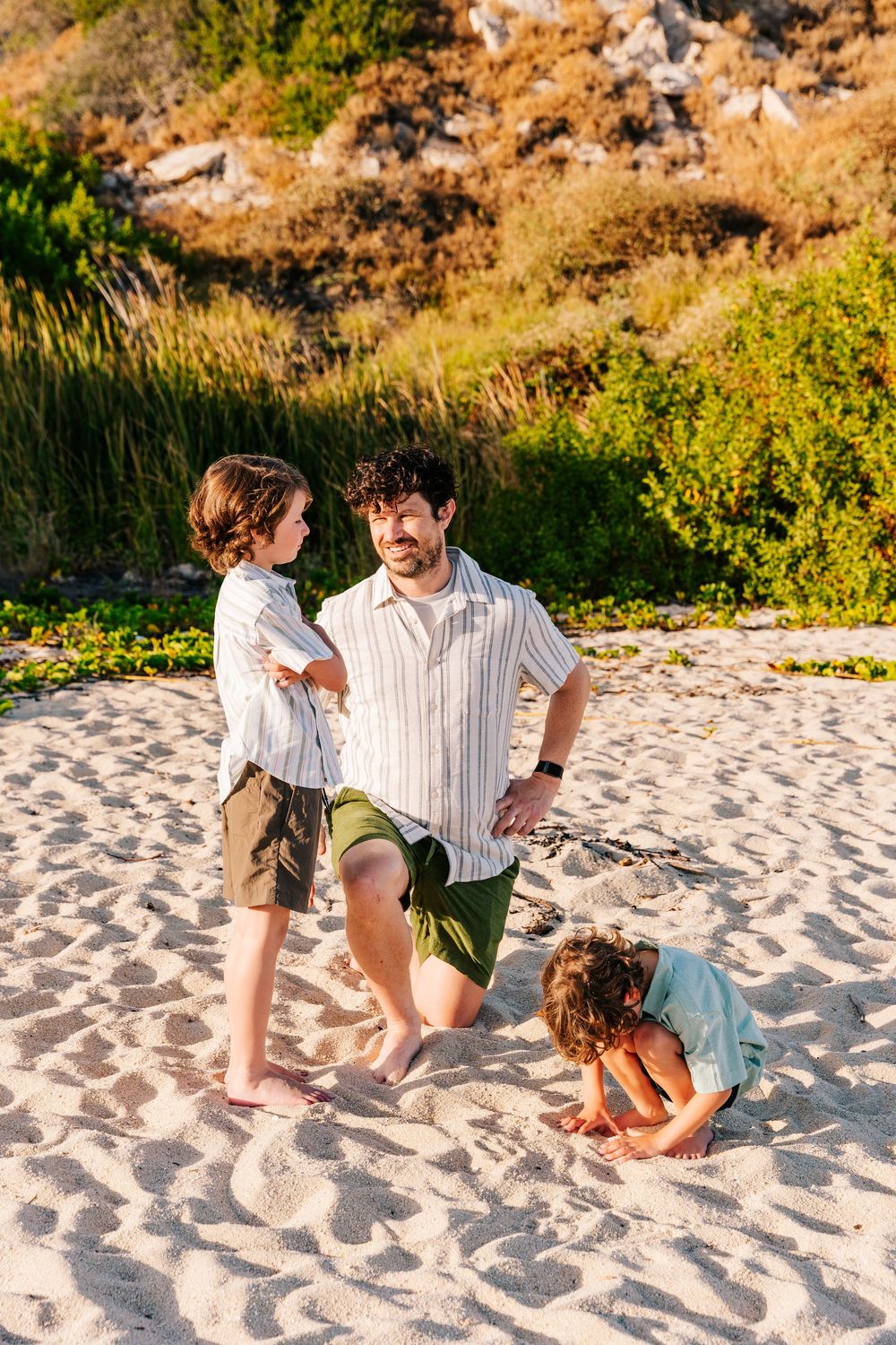 Father kneeling on sandy beach playing with two young children, surrounded by lush green vegetation in warm sunlight.
