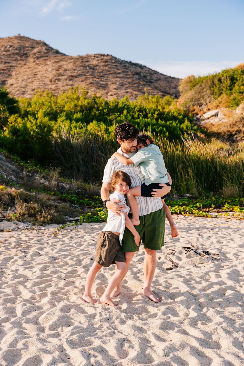 Father hugging two young children on a sandy beach with green hills and blue sky in the background.